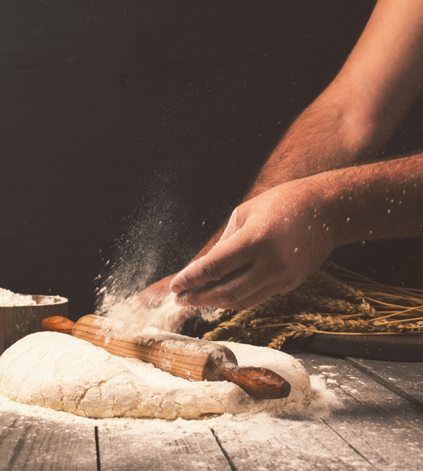 pizza dough on a table with a rolling pin in the middle, hands tossing flour onto it